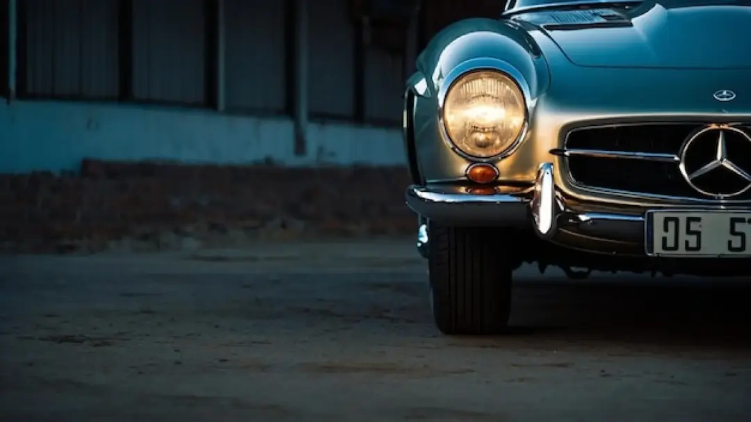 Classic silver Mercedes-Benz with its headlamp on, parked outside a building at dusk